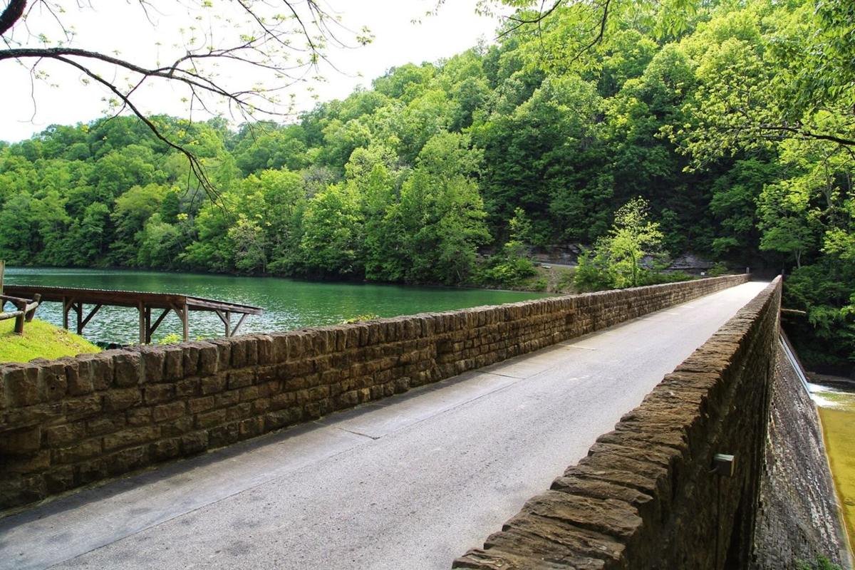 This image shows a 1 lane bridge and dam in the Standing Stone State Park. A row of rental paddle boats exist in the left side of the picture, and the road meanders up a hill to a scenic overlook.