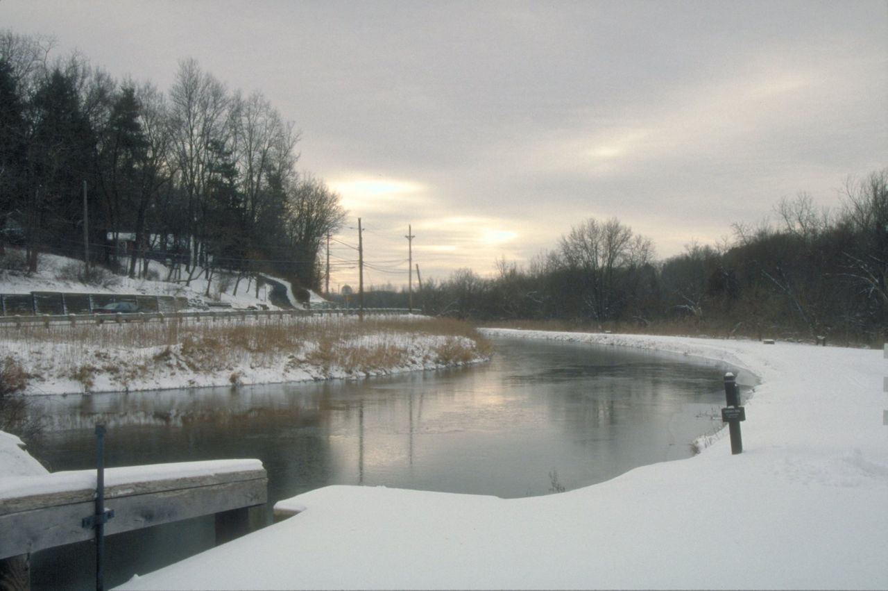 A canal behind the Canal Visitor Center in Cuyahoga Valley National Park is partially iced over during the winter. There is snow on the ground, and the remote sun is setting.