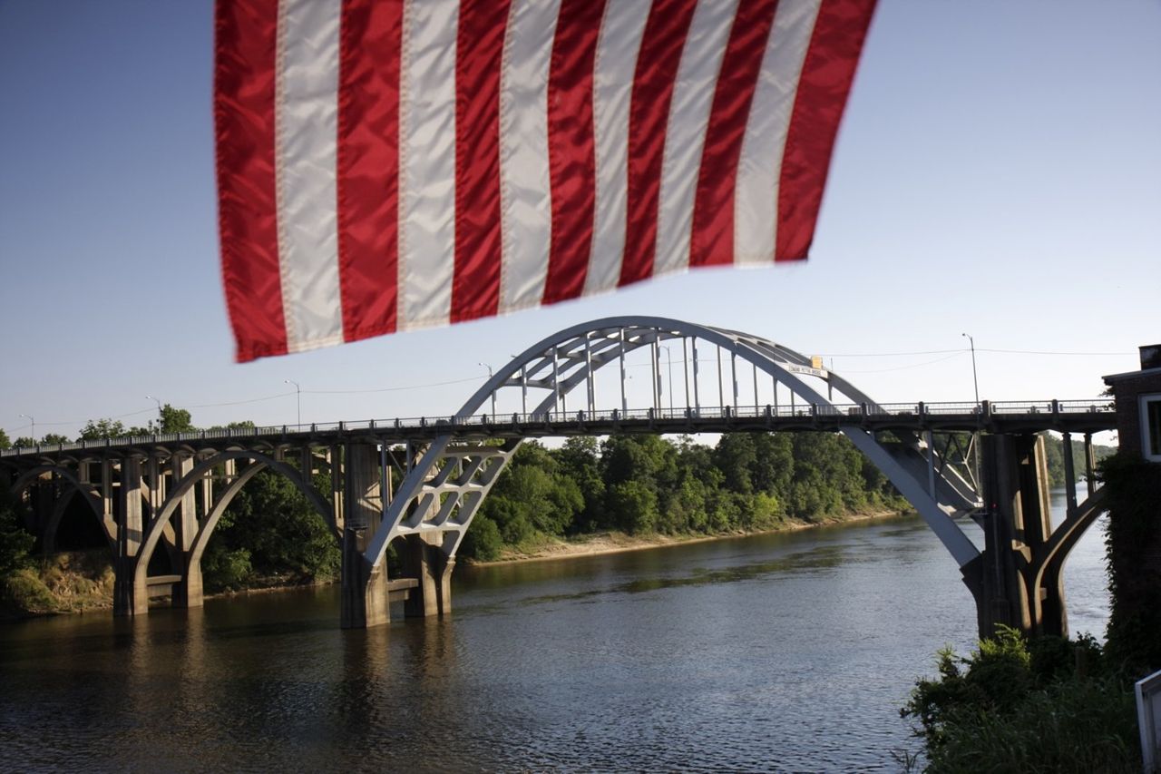 The red and white stripes of the American flag hang over a view of the Edmund Pettus Bridge crossing the Alabama River in the distance.